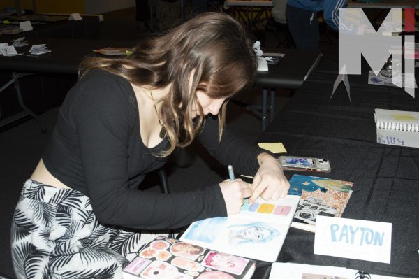 Writing on a sticky note, senior Dory Latenser compliments a fellow artists sketchbook collection. It was Latensers first time at the event and displayed  over 10 pieces. 