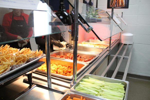 Setting out trays of wings, mac and cheese and veggies, cafeteria workers supply warm meals for lunch. Split between the opposite side of the kitchen, alternative food options are provided. 